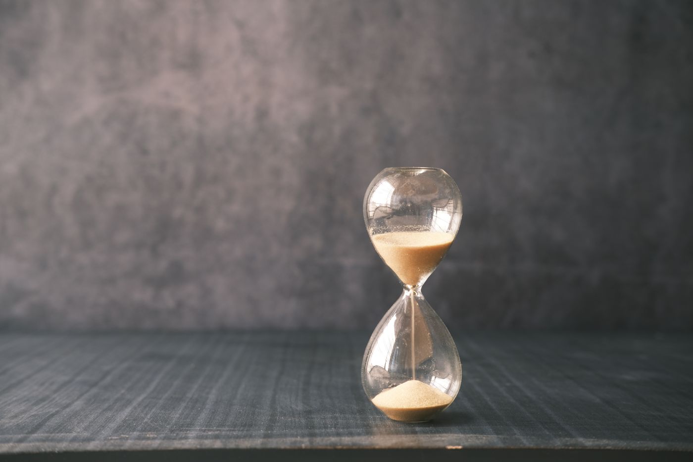 A glass hourglass standing on a dark wooden surface against a grey wall, its sand part way through the neck.