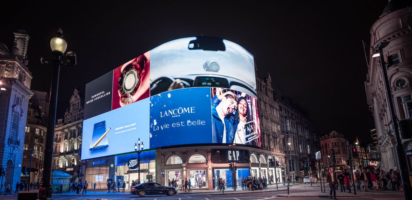 Piccadilly Circus in London at night, its curved digital billboard lit with advertisements for Lancôme, Samsung and others above the Boots and Gap shopfronts, with people on the pavement below.