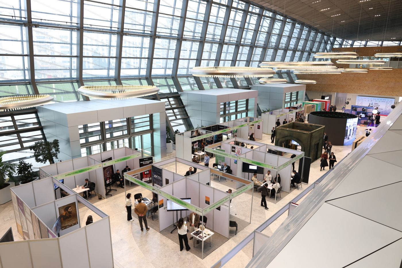 An exhibition hall seen from above: rows of white modular stands under a tall glass wall, with small groups of people talking at tables between them.