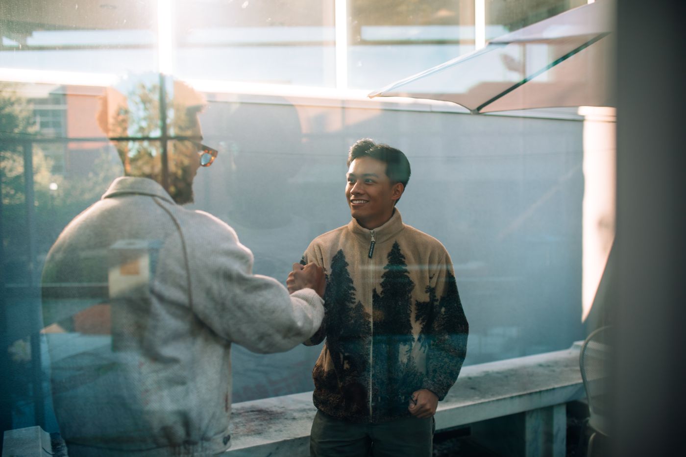 Two men shaking hands beside a sunlit window, photographed through the glass of the office next door.