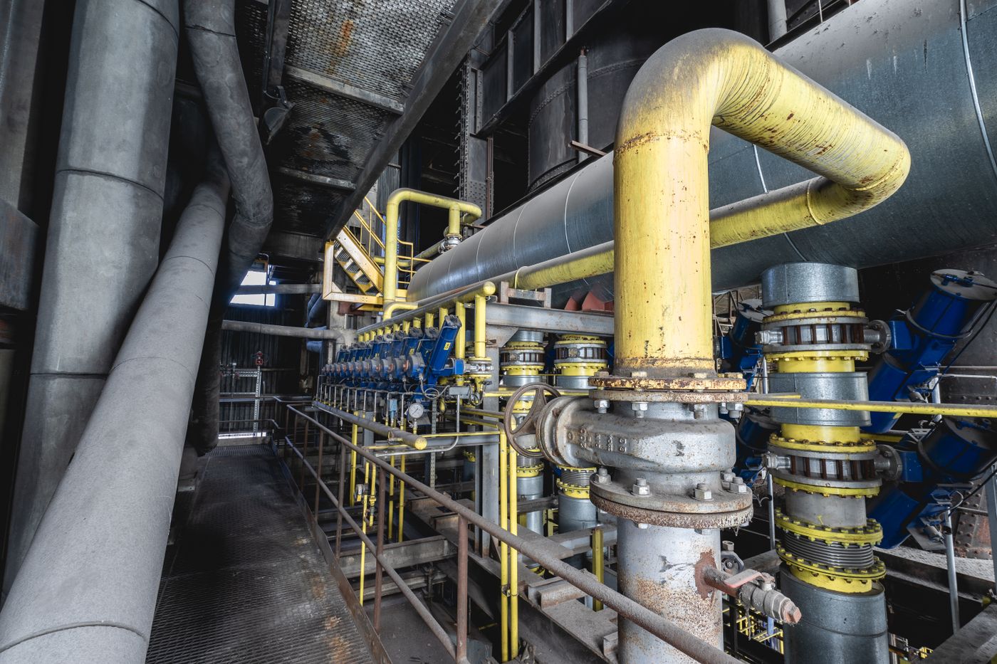 A yellow gas main curving down into a row of flanged valves and branch pipes inside a disused industrial plant.