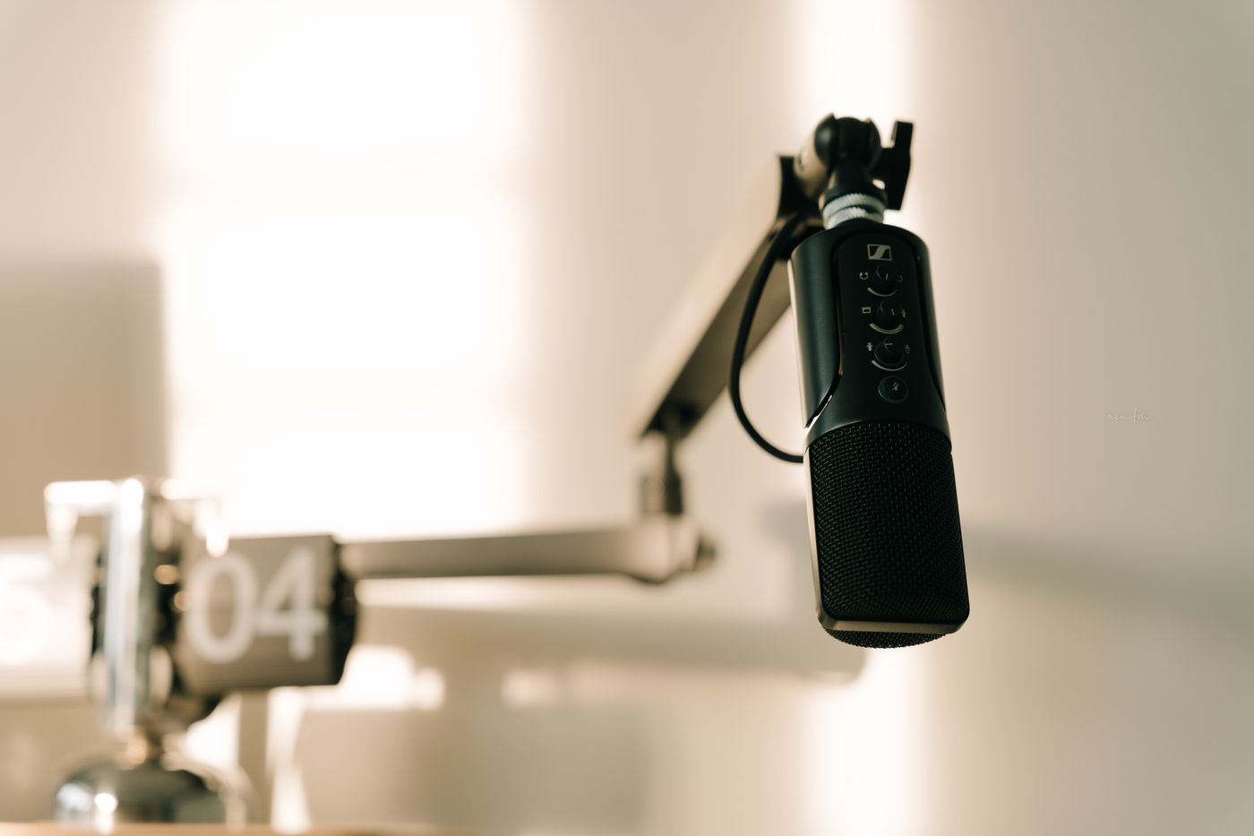 A black broadcast microphone on an articulated boom arm in a softly lit room, with a flip clock reading 04 out of focus behind it.