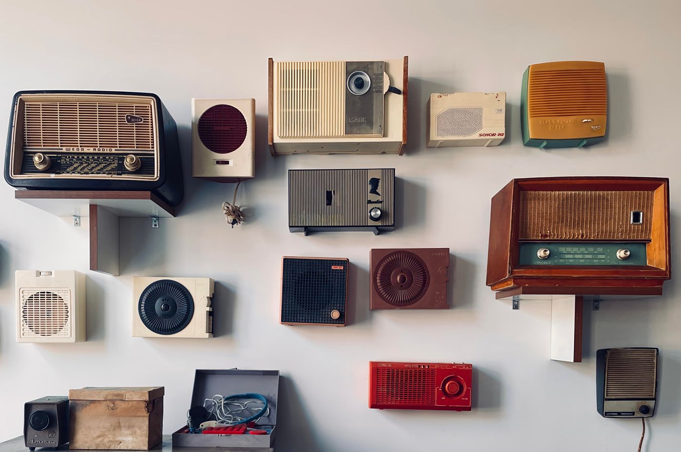 A white wall hung with more than a dozen vintage radios and speakers on small shelves, from a black Wega tabletop set to a wooden console radio and a red portable.
