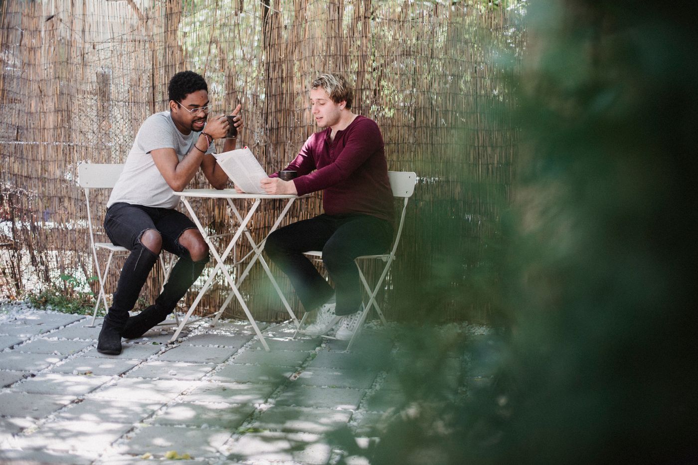 Two men talking at a small table in a sunny courtyard, one holding up a newspaper.