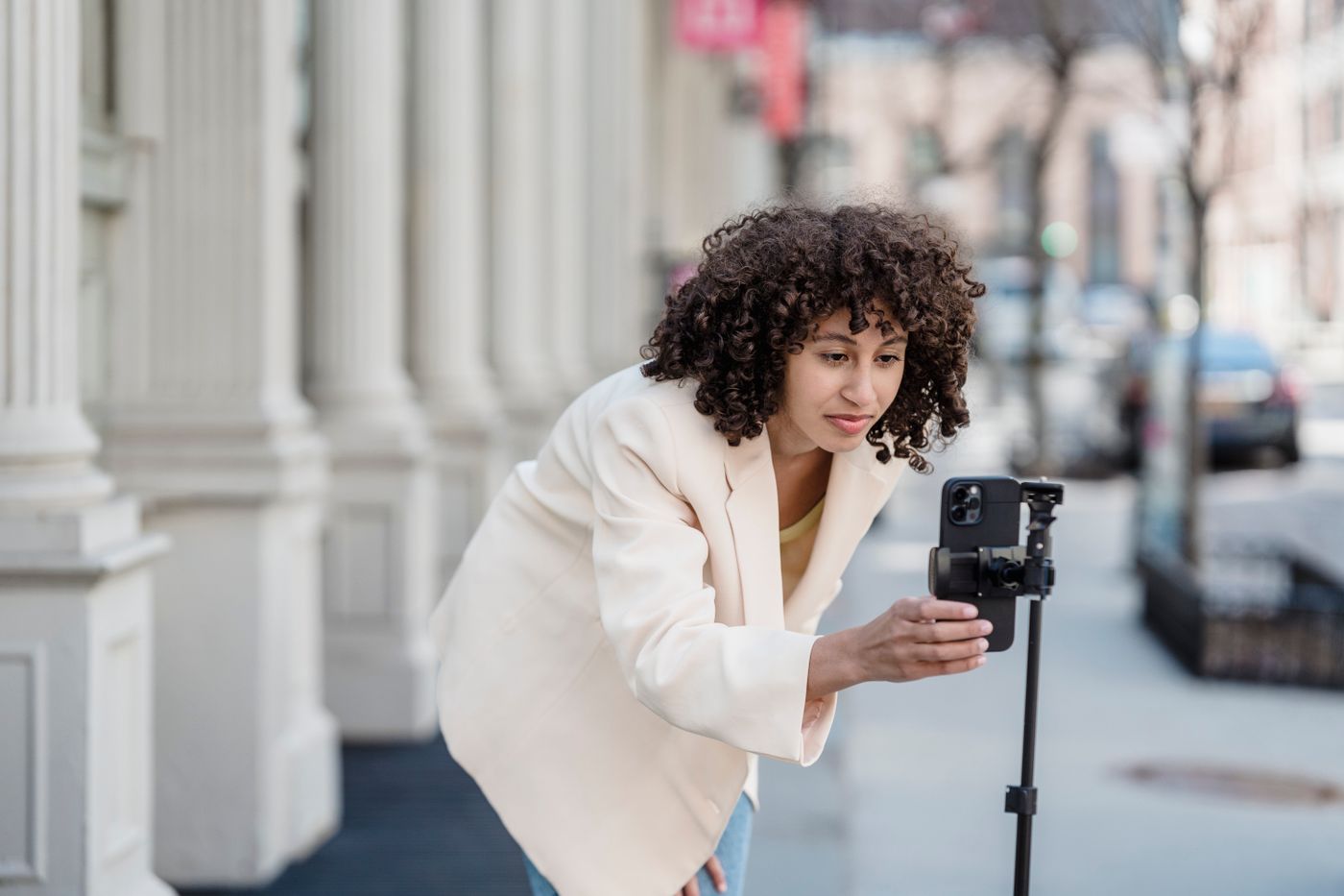 A woman on a city sidewalk leaning in to frame a shot on a smartphone mounted on a small tripod.