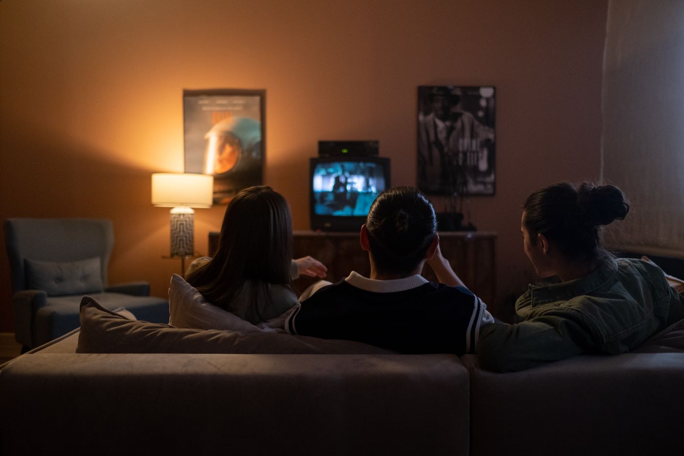 Three people on a sofa, seen from behind, watching a television in a lamplit room.
