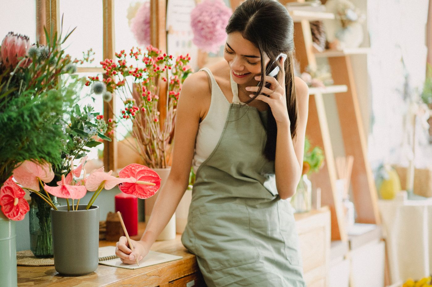 A smiling florist in an apron talks on her phone and takes a note at the counter of her flower shop.